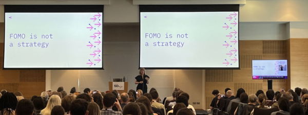 Rachael presenting at the SDGov conference. She stands in the middle of a room, facing an audience. Besides her, are two large projection screens with the quote "Fomo is not a strategy" displayed on both of them.