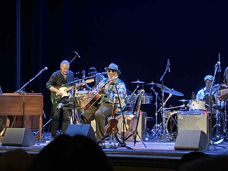 An elderly man in a fedora and sunglasses sits on a stool onstage holding an acoustic guitar. Slightly behind him to his right is a man playing a Fender Telecaster.