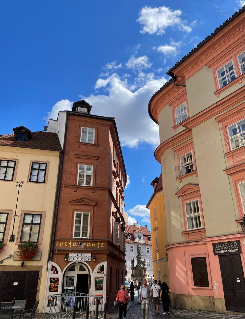 A narrow street opening sandwiched between two buildings. The one on the left has a brown large smooth brick like facade and has a restaurant on the ground floor. The one on the right has a pink and cream colour pattern with old windows and curves on one side. At the far end of the opening, there is a statue in a small neighbourhood square. There are a few tourists walking in the foreground