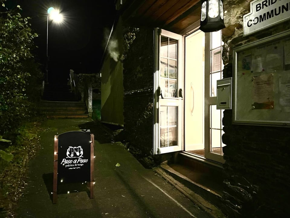 Photo of the entrance of the Bridgetown Community Hall in the evening. The window door is open, with a lantern hangin from the porch roof and a blackboard pavement sign of "Paso a Paso" in front of the door. On the wall, to the left of the door, the sign and of the Bridgetown Community Hallo is slightly visible. In the background, on the centre-left, the four steps connecting the hall to the above parking lot "