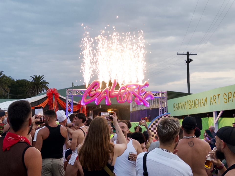 A lively crowd gathers at the entrance to Tropical Fruits, with people taking photos of a pink neon “Galore” sign on a stage, illuminated by a burst of fireworks overhead.