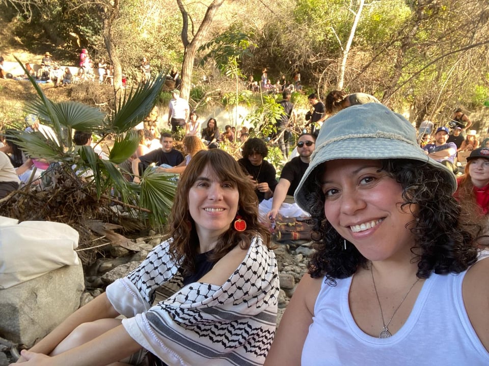 A smiling selfie of Lauren and Xochitl in the riverbed, surrounded by people, river rocks, and sunlit trees. Xochitl is rocking a bucket hat and curly hair over a white tanktop. Lauren is wearing dangling red earrings in the shape of cut pomegranates and a black and white keffiyeh draped over her shoulders.