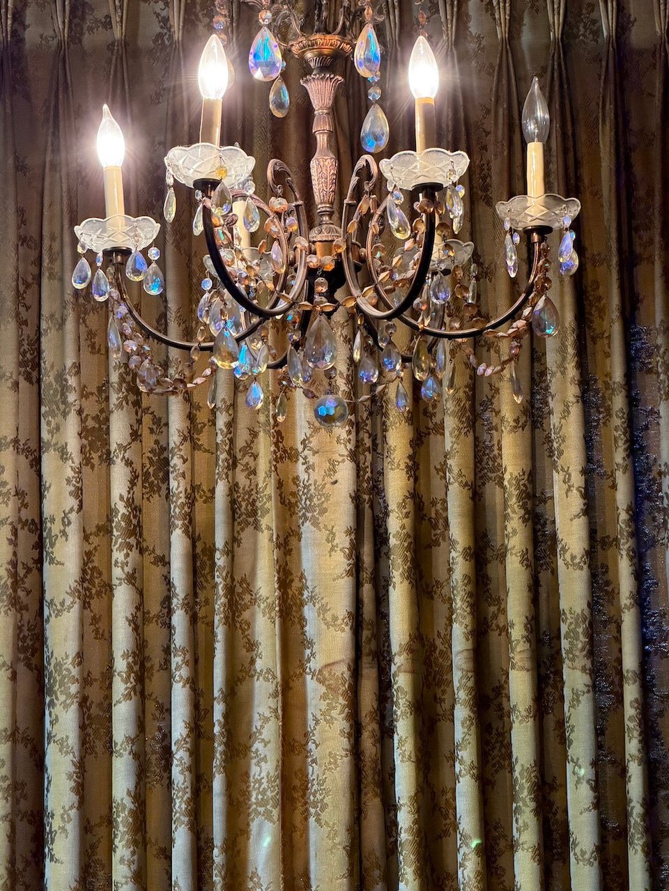 a chintzy crystal chandelier with one bulb out in front of aged formerly golden drapes