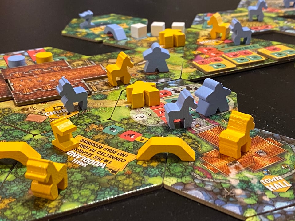 Tiles with pictures of trees, paths, and houses put together to form a village. Wooden brides, meeples, and donkeys are on top of the tiles.
