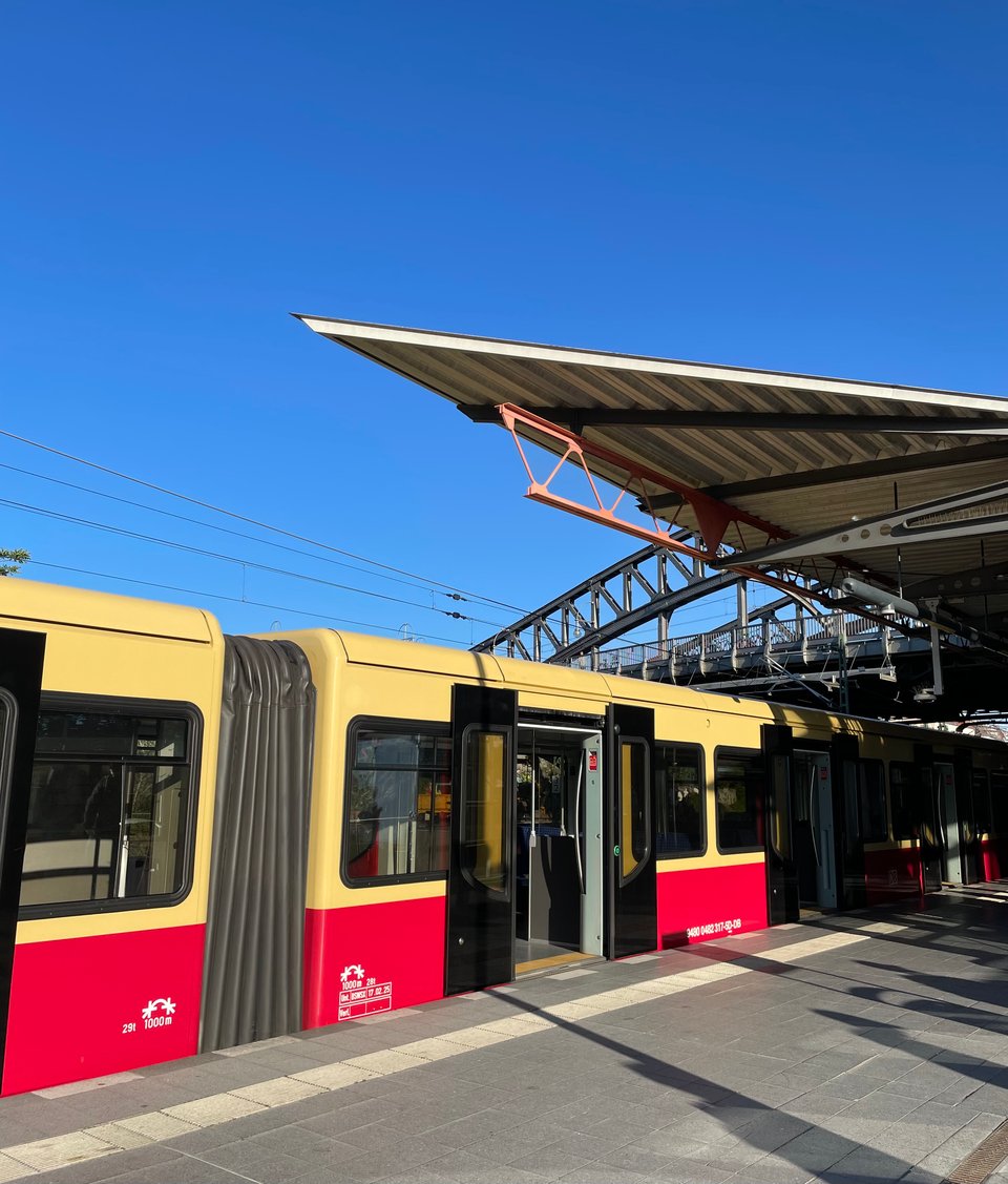 A view of the S-Bahn train at a station in the evening. There is the red and yellow train, an overhead platform shade ending in a pointed edge and long shadows on the platform