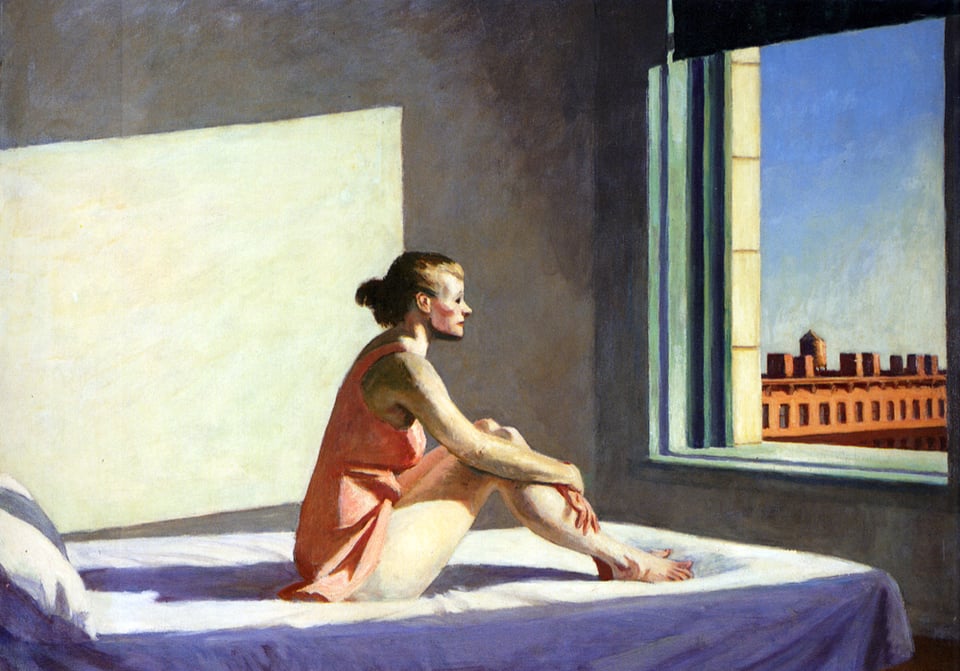 A woman in a pink slip sits on a bed in a sunlit room, gazing out of a window toward a brick building across the street.