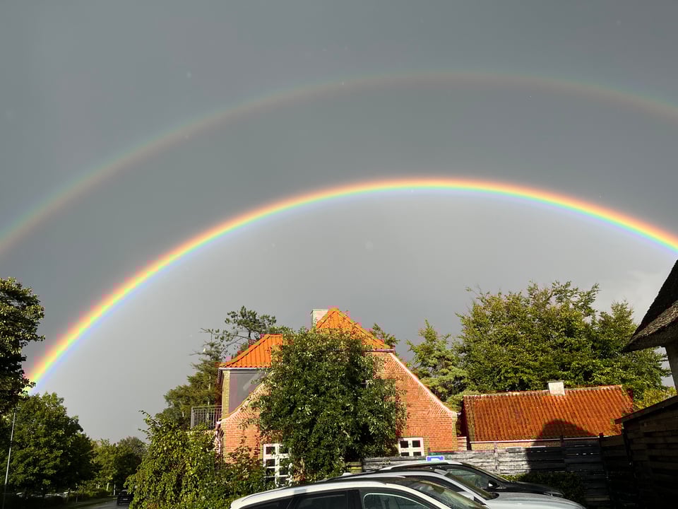 Above a brick house with orange tiled roof span two rainbows, the upper rainbow pale and the lower rainbow deeply intense.