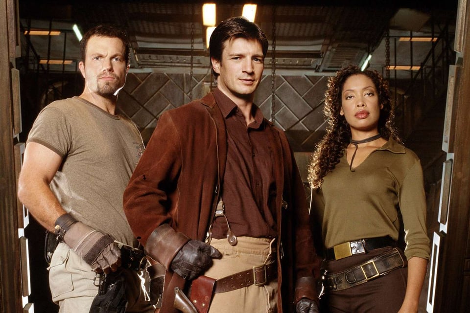At left, Adam Baldwin, a bearded white man, wears a t-shirt and gloves and scowls at the camera. In the center, Nathan Fillion wears his trademark brown coat and reddish brown shirt while showing off his suspenders and gunbelt. At right, Gina Torres wears a green shirt, brown pants and visible gunbelt. Only Fillion is smiling.