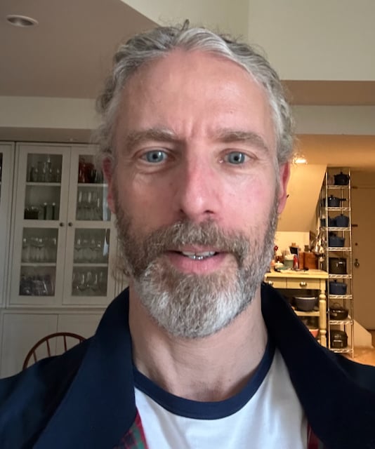 Indoor photo of a white man in a white ringer T-shirt with a blue collar, and a navy blue jacket.