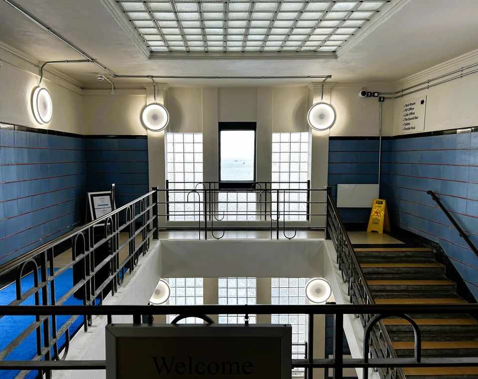 The same stairwell without any people but with a 'wet floor' warning sign. plaster has been restored and large circular lights fitted. The tiles and floor can now be seen in their original 1930s colours of blue, red and black.