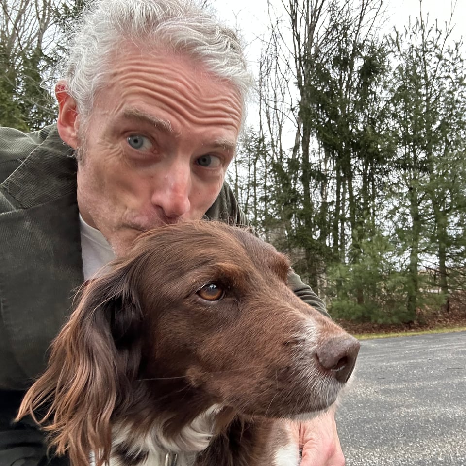 Photo of white man smooching the back of a brown dog's head; the dog looks like a spaniel/terrier sorta mix