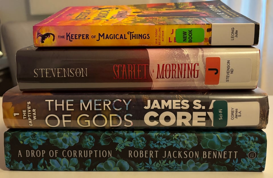Stack of books, spines out: The Keeper of Magical Things bu Julie Leong, Scarlet Morning by N.D. Stevenson, The Mercy of Gods by James S.A. Corey, and A Drop of Corruption by Robert Jackson Bennett