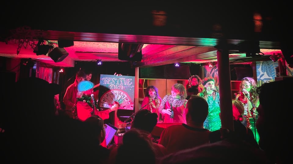 8 musicians crammed onto a stage in festive outfits, lit in bright pink, red, and green