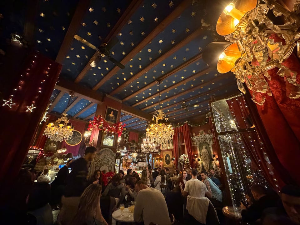 A bar is full of people sitting down and lights and chandeliers throughout.