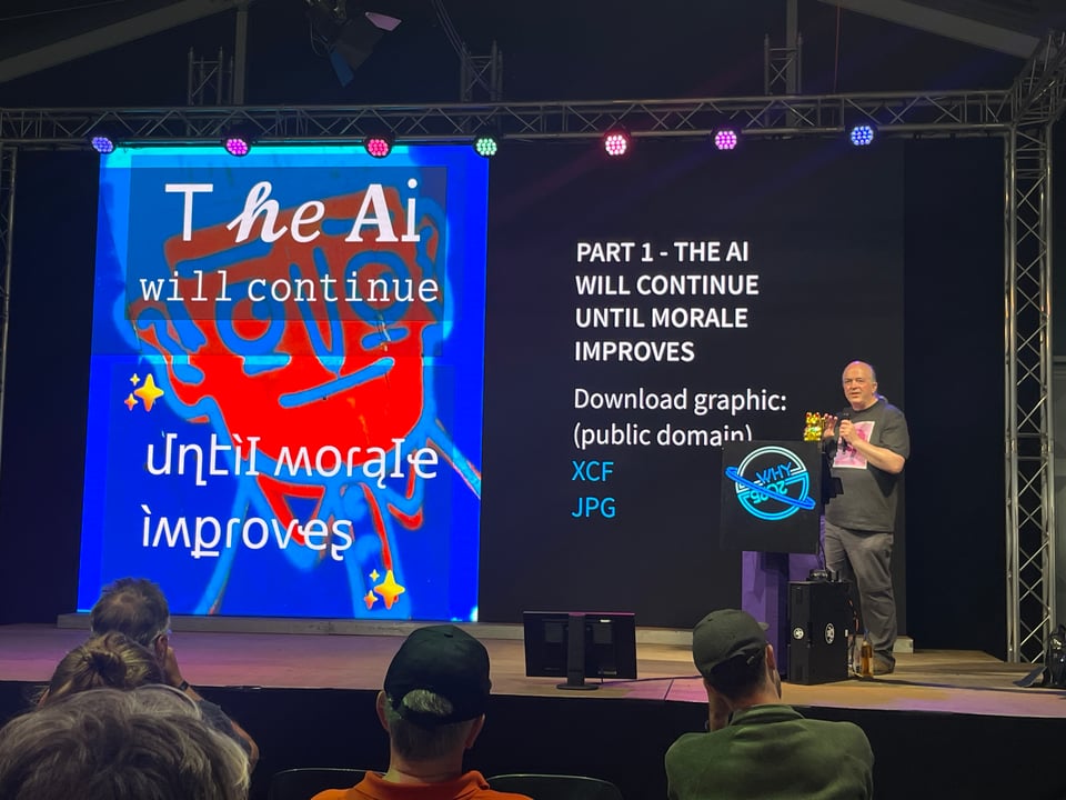 A man speaks on stage with a WHY2025 logo on the podium. Behind him the screen reads “The AI will continue until the morale improves.”