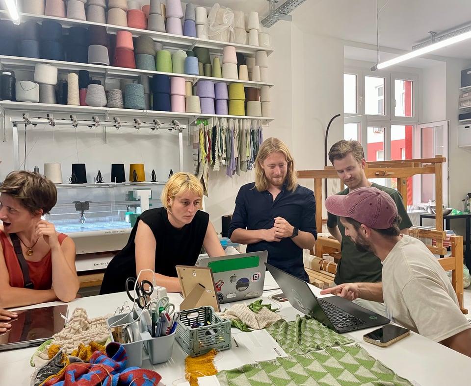 People centred around a table with many fabrics, tools for textile work and laptops. Creating designs that the machine in the background of the photo will print. The wall against which the machine stands, has multiple shelves of colourful yarn on it