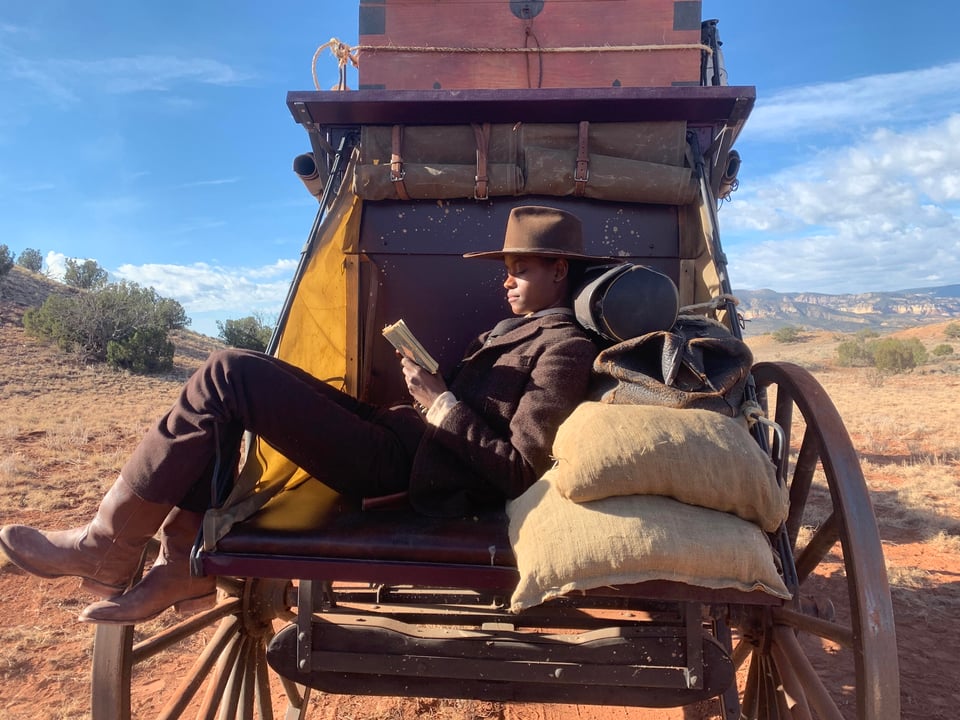 A Black person sits on the back of an old Western wagon and reads a book