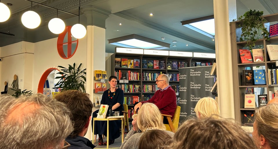 A man and woman sit talking to each other in a raised platform in a bookshop. A small audience listens to them.