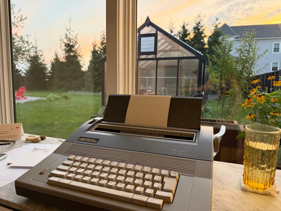 Picture of a grey and blue typewriter sitting on a marble top desk, with a full glass of water in a textured yellow glass. Through the window there is a cluster of flowers, and a greenhouse, with a hint of a light gray house in the distance, and a blurry row of short evergreen trees.