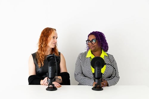 Nicole and Patricia sitting behind microphones and looking at each other dubiously