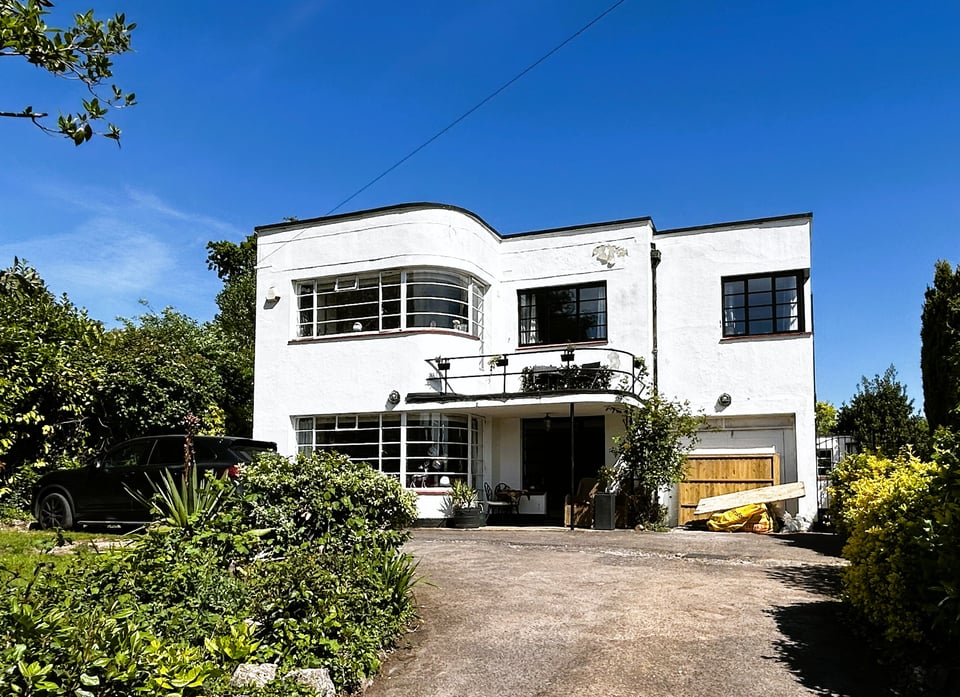 A detached house up a driveway. To the left are two curved windows of Crittall glass. A small balcony is on top of the porch, also with a curve. There's an integral garage on the right.