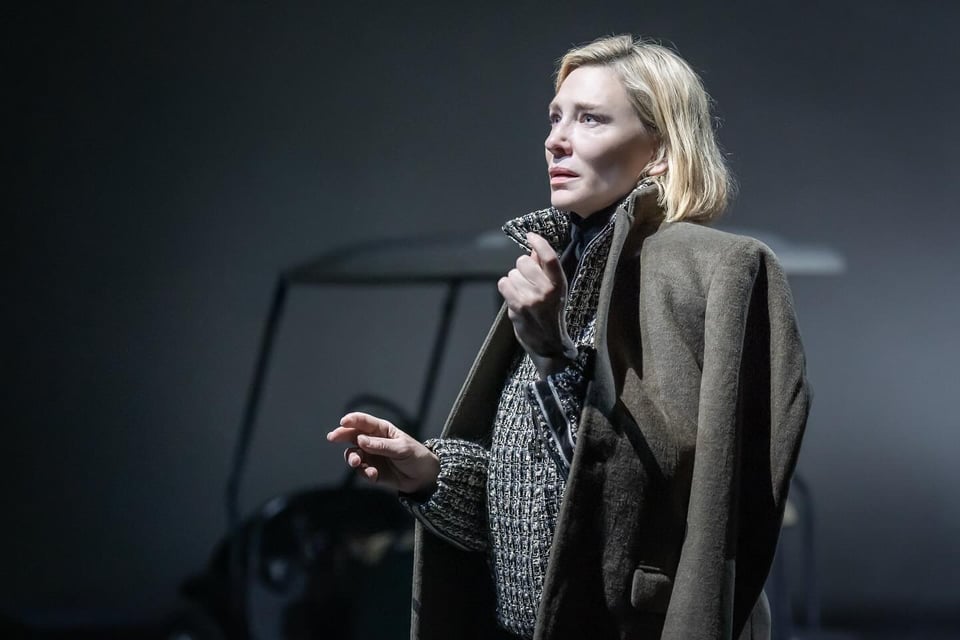 An image of Cate Blanchett in Thomas Ostermeier and Duncan Macmillan's adaptation of The Seagull. Blanchett has blonde hair and is wearing a big grey coat and a grey knitted fleece. She is standing in front of a golf cart.