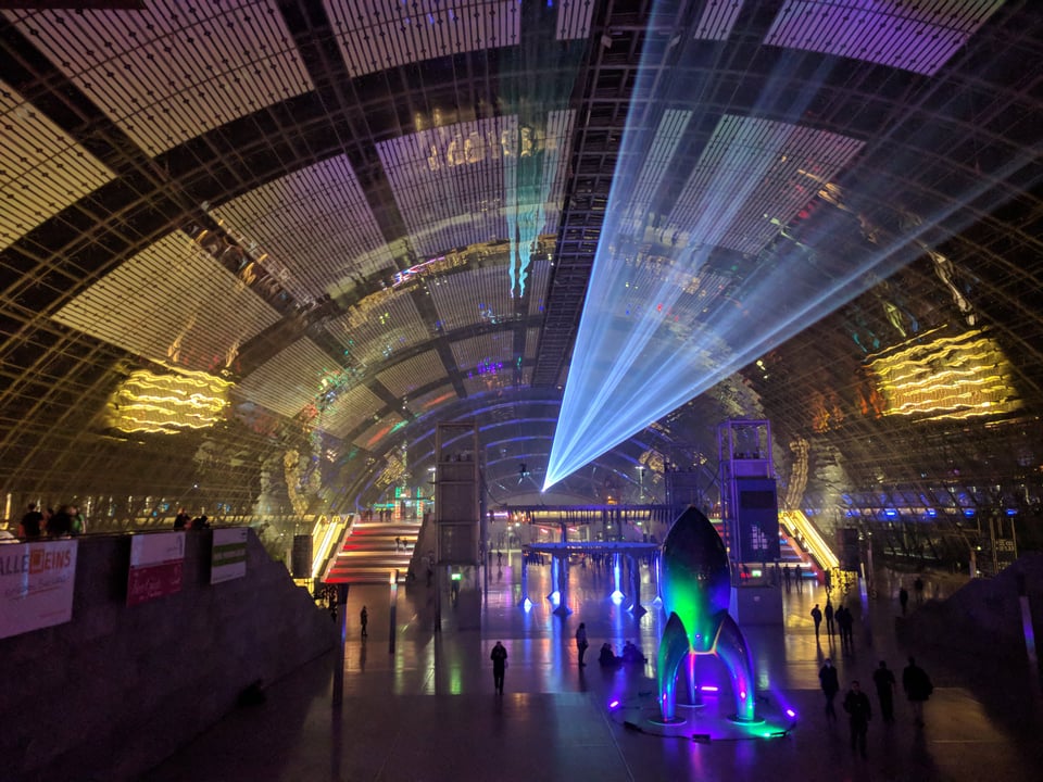 A large conference hall at night with a blue laser shooting across the glass ceiling and a large model rocket lit with green, blue, and purple lights.