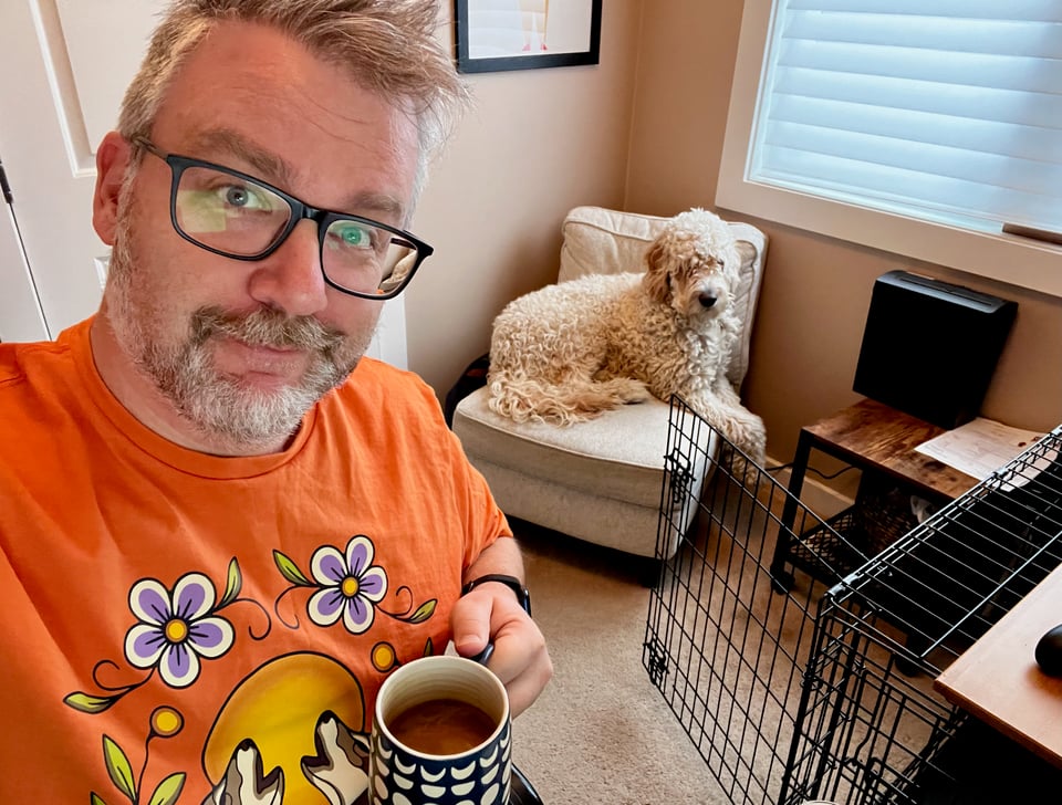 Me sitting with a mug of coffee in my hand looking at the camera. Our dog, Neville, sits on a chair behind me. He barely fits on it.