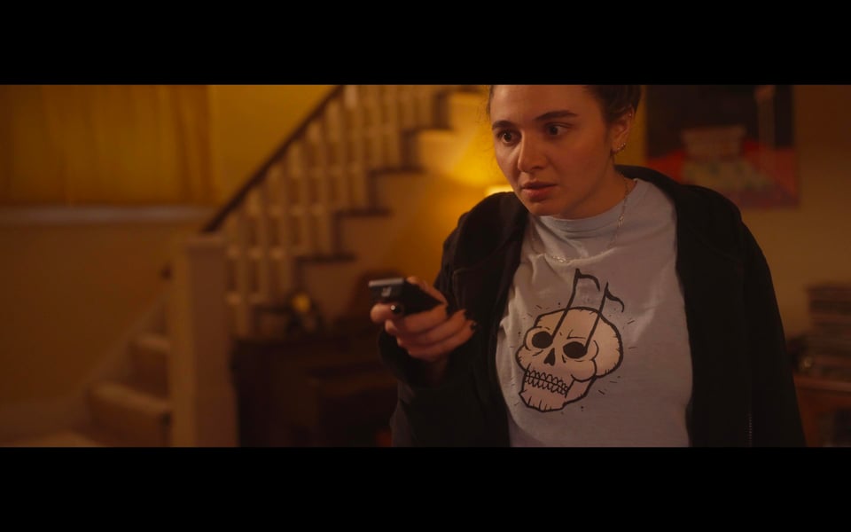 A screenshot from a horror film. A young woman stares intently with a TV remote in her hand. She wears a t-shirt with a skull who has music notes for eyes
