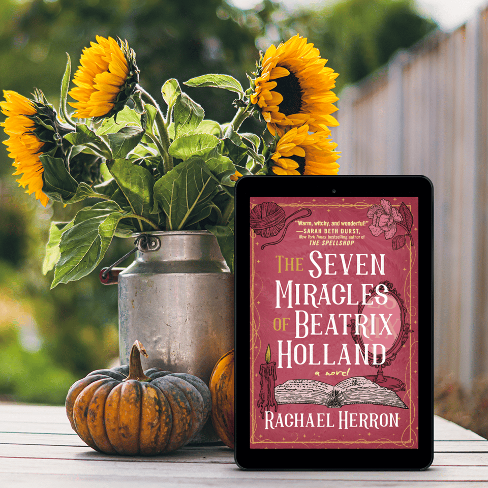 pink book, The 7 Miracles of Beatrix Holland, on a table with yellow daisies and orange squashes