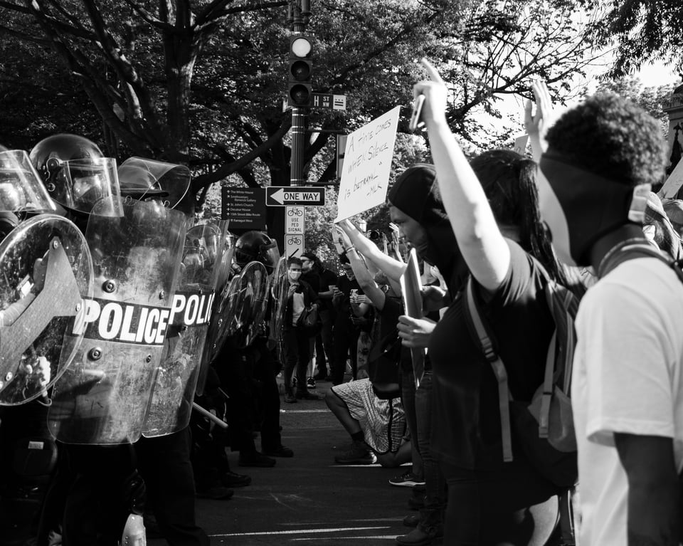 Protesters face a police line. In the background is a "One Way" sign, pointing in the direction the protestors are going, indicating the police would have to move back. One of the protestors holds a sign "A time comes when silence is betrayal." a quote attributed to MLK, but was actually him quoting a letter from a group of clergy about the Vietnam War.