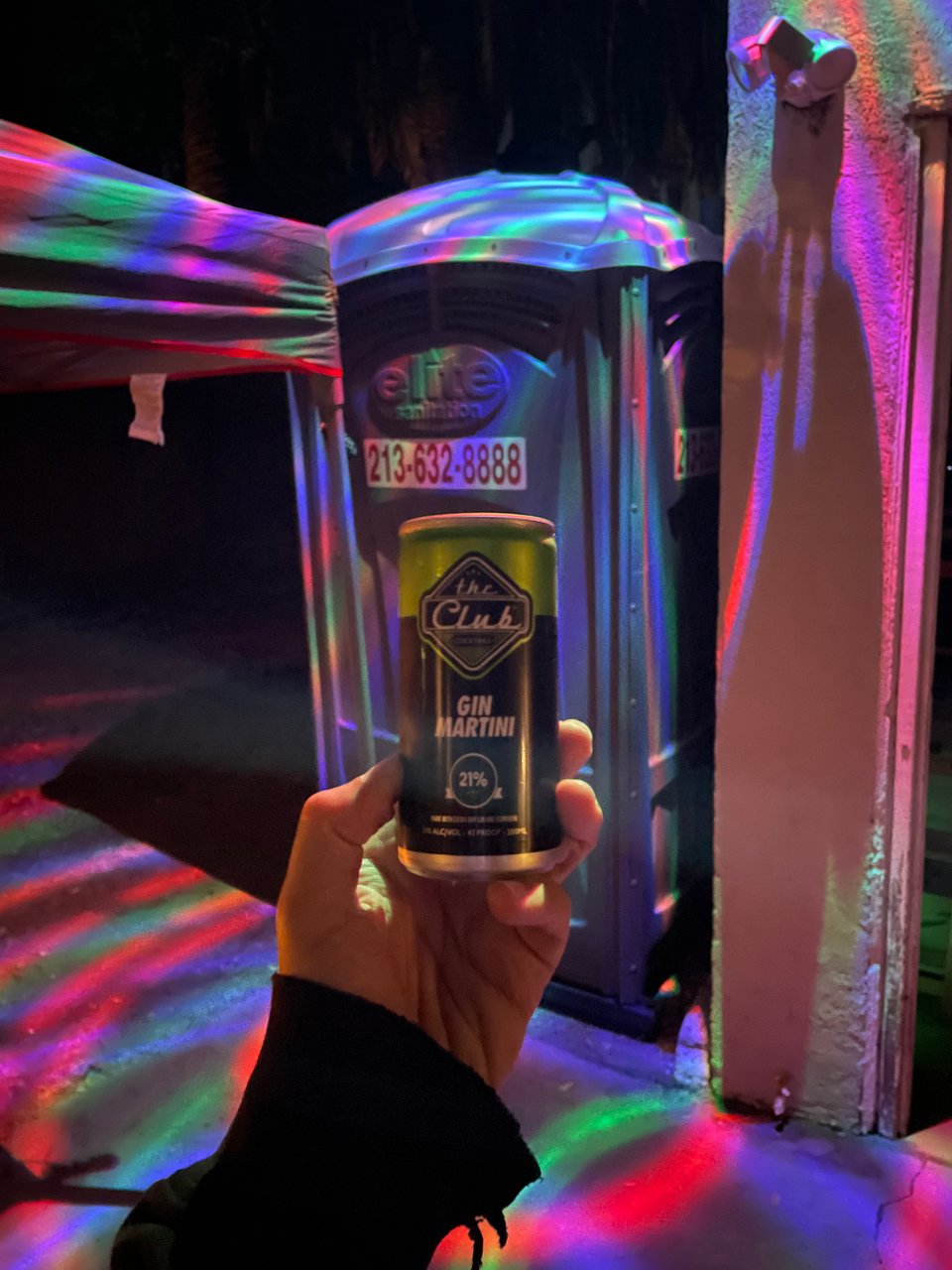A hand holds a gin martini in a can in front of a port-a-potty bathed in spinning dance floor lights