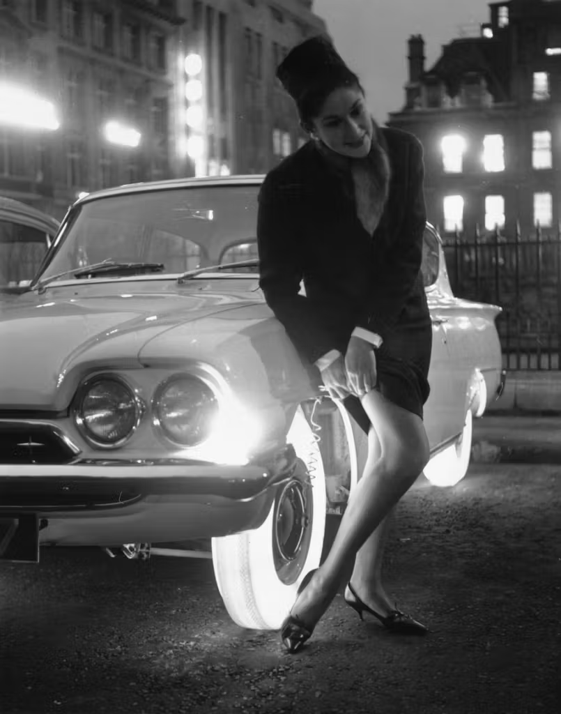 A black-and-white photo of an old-fashioned car from the 1950s, with the tires brightly glowing white. A woman in a tight dress with long sleeves and a pillbox hat is standing directly next to the car's front lefthand tire, and she is reaching down to slightly pull her skirt up over her right knee, as if she were adjusting something in her clothing. The back of her knee and legs are illuminated brightly by the glowing tires. In the background are several large, tall, old-fashioned, multi buildings. The effect is very dramatic.