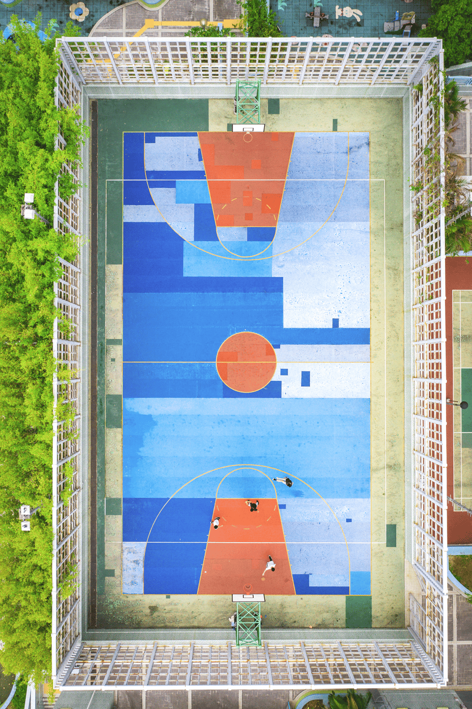 An aerial picture of a large basketball court. It is very colorful and divide it into different geometric shapes, including dark blue triangles, light blue rectangles, and burnt orange circles and parallelograms. The is surrounded by a high white fence and has a forest of green trees on the left side and what looks like another smaller court on the right side. There are four people playing basketball, one is shooting the ball towards the hoop