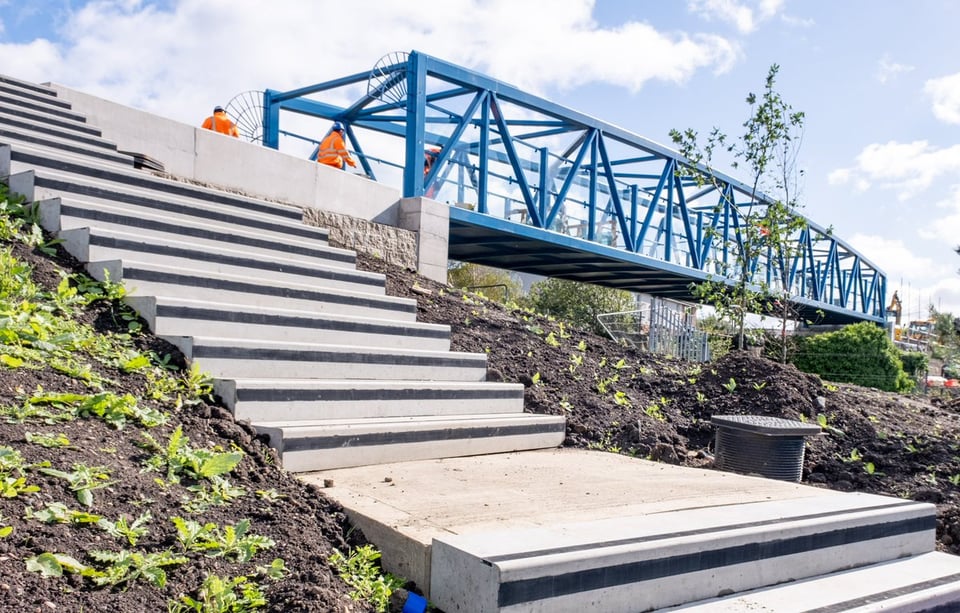 Three people in bright orange hi-visibility jackets work on a pristine blue iron bridge span, while in the foreground multiple flights of new concrete steps are seen leading down muddy banks and new planting to Russell Road.
