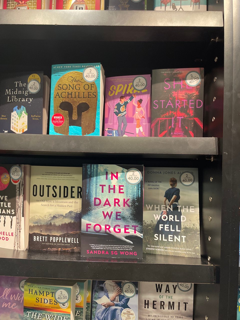 Photo of a bookshelf at Vancouver International Airport with my novel IN THE DARK WE FORGET among others such as SONG OF ACHILLES, FIVE LITTLE INDIANS, and THE MIDNIGHT LIBRARY, all with stickers that read “2 for $40”