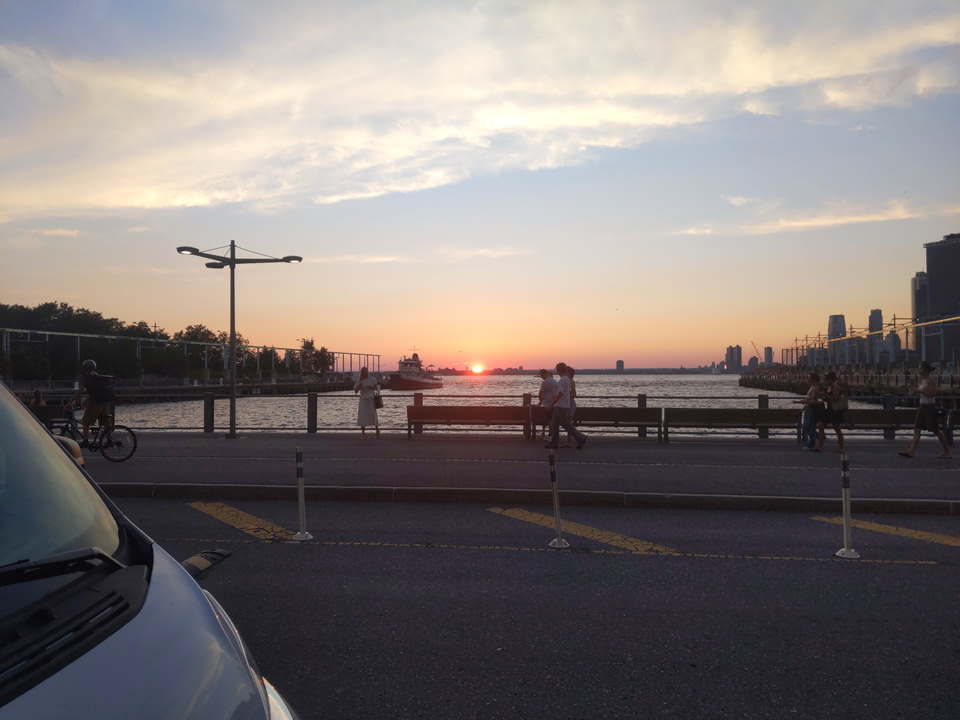 Across the street from the water in Brooklyn Bridge Park, we see the setting sun off in the distance beyond New Jersey. In the immediate foreground is a car. We also see two piers (framing the Sun) and a boat off in the distance.
