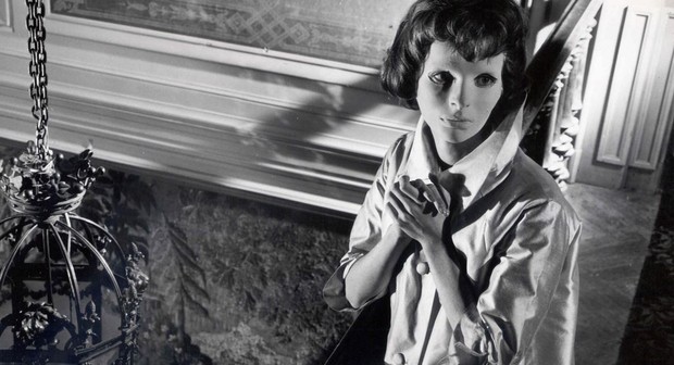 Edith Scob, wearing an expressionless mask showing only her eyes, stands at the side of a room with her hands clasped, in Eyes Without a Face.