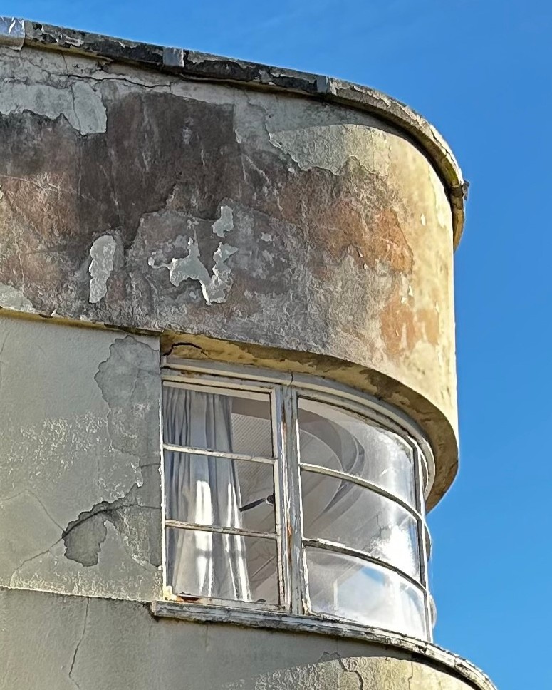 Close up of a curved window set back from cream rendered walls. The metal window frames are rusting and bubbled, but the glass is original. The render itself is mottled with damp, cracked and in some parts missing.