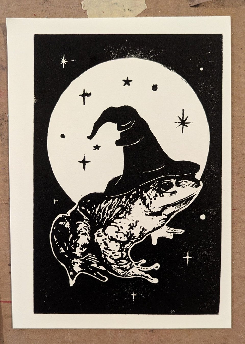 A black and white illustration of a frog wearing a witch hat against a large moon.