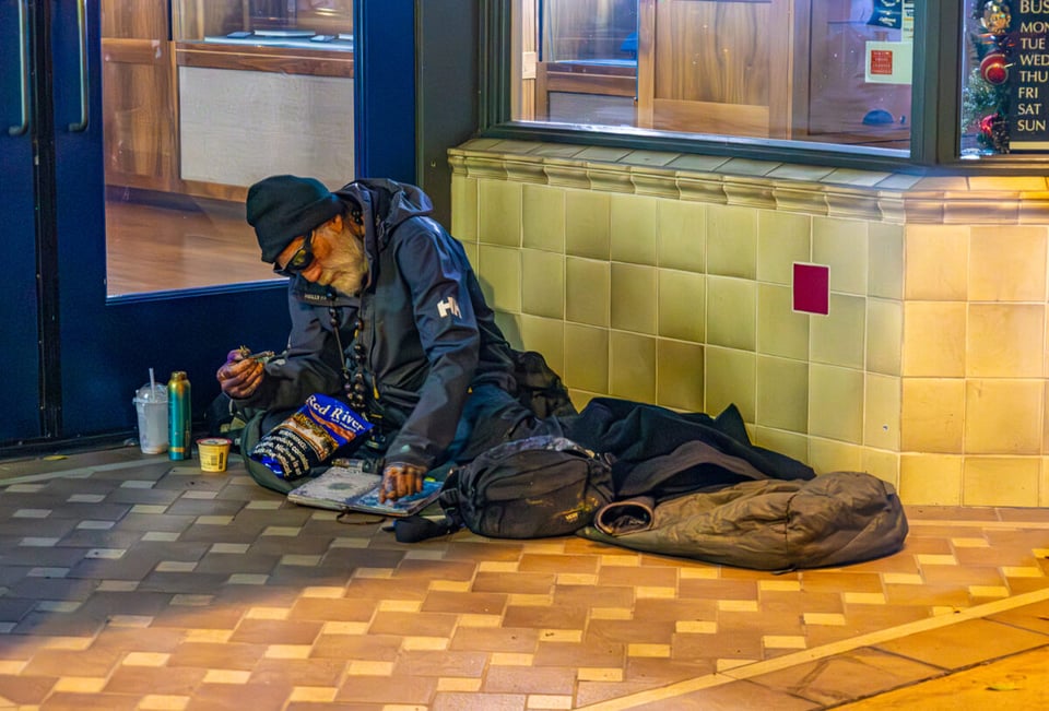Painting of a sleeping unhoused man with white beard wearing sunglasses laying propped up in an entryway to a store. Around him is a half drank boba tea, a thermos, a bag of chips, a notebook, his backpack, and a sleeping bag.