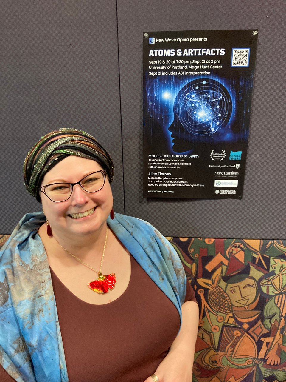 A white woman with glasses wearing a headwrap, a blue scarf, and a burgundy dress simles in front of a poster for New Wave Opera's performance