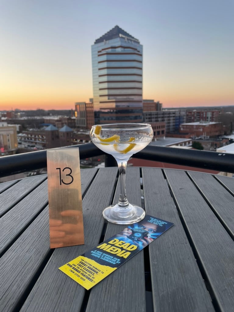 A martini, a promotional bookmark for Dead Media, and a golden stand with the number 13 on it all rest on a table. Behind them is a tall building and a vista of a city in the setting sun