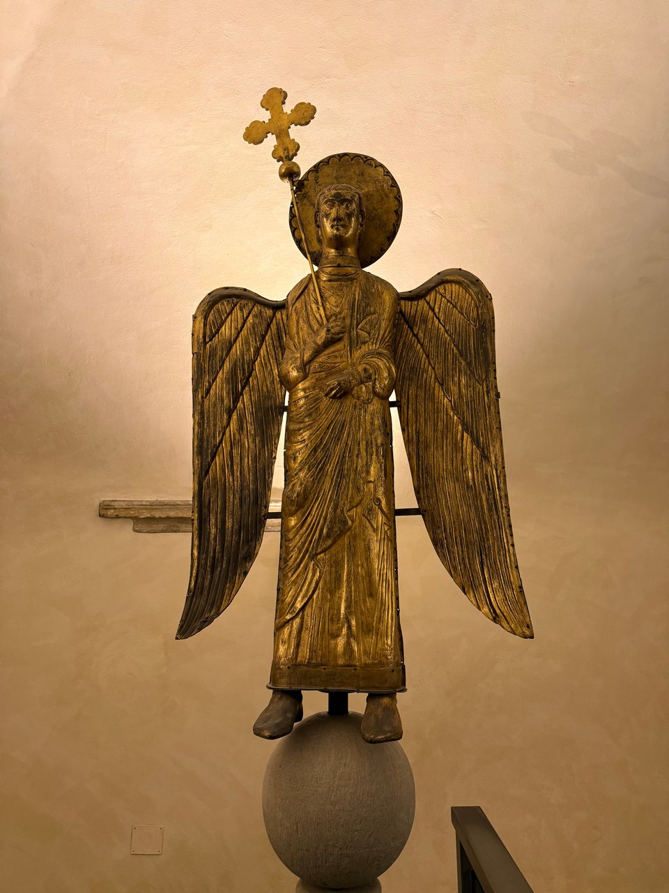 Photo of copper angel statue from post-12th century Parma. It is holding a staff w/a cross, and is standing on a globe. There is a disc behind its head to represent a halo.