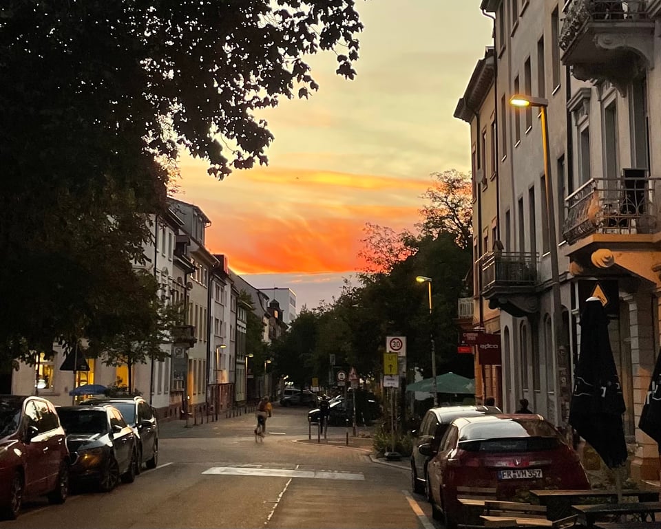 A view of the street sloping down with parked cars on both side. A part of a tree hanging on the top left and a magnificent orange sunset sky in the distance. The shades of orange go from almost red to almost yellow, all reflected on the clouds in the sky
