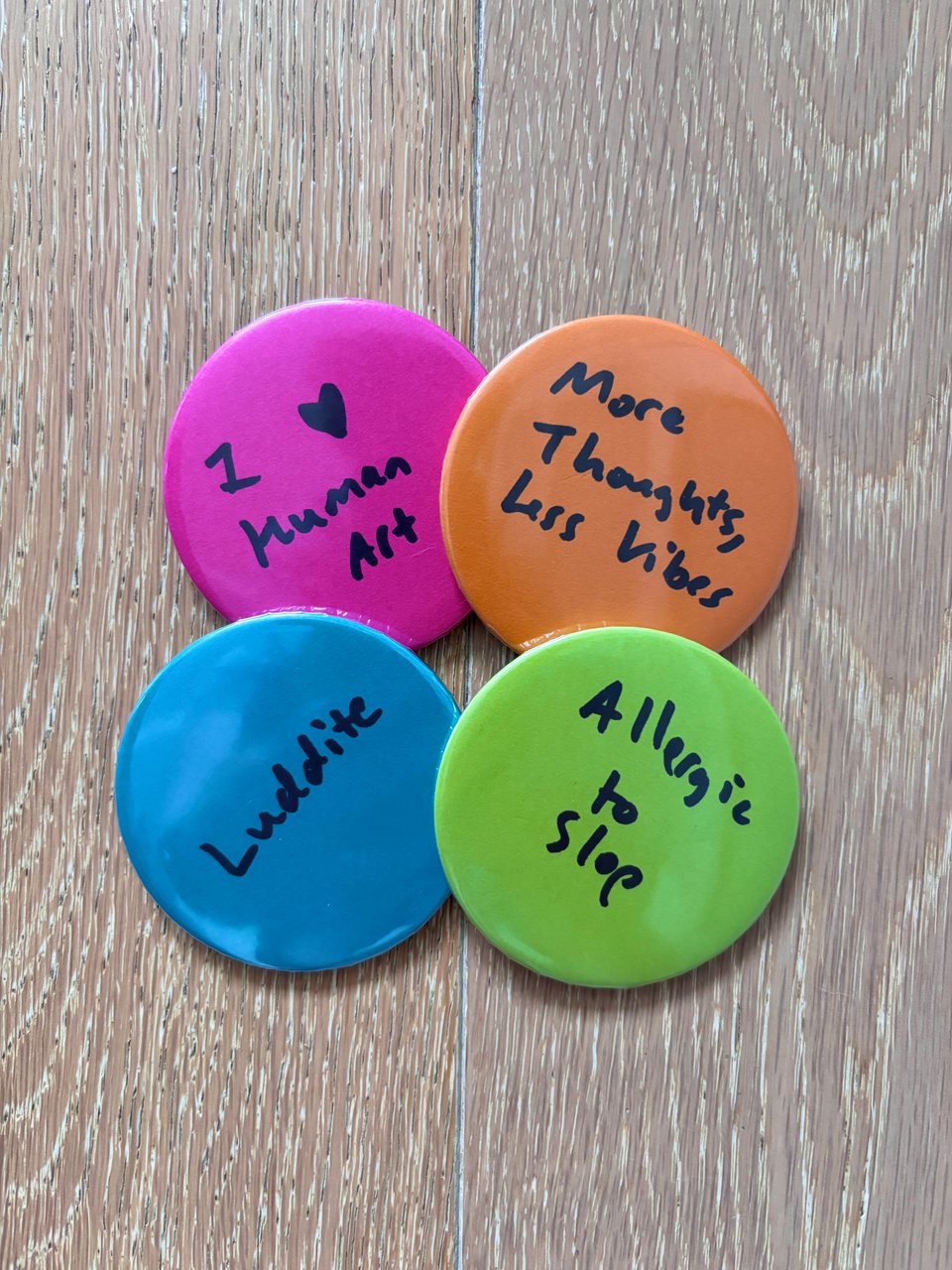 Four button pins arrayed in a square, all with handwritten text in black ink. Top left: pink pin that says "I love human art". Top right: orange pin that says "More thoughts, less vibes". Bottom left: blue pin that says "Luddite". Bottom right: lime green pin that says "Allergic to slop"
