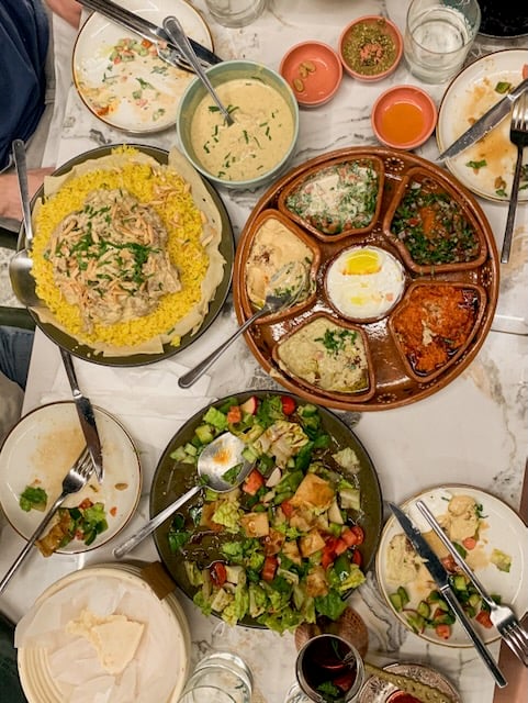 A spread of salad, mezes, and lamb served over rice sits on a white tablecloth