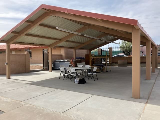 A metal-roofed pavilion with open sides over a concrete patio with shaded plastic chairs and a table.