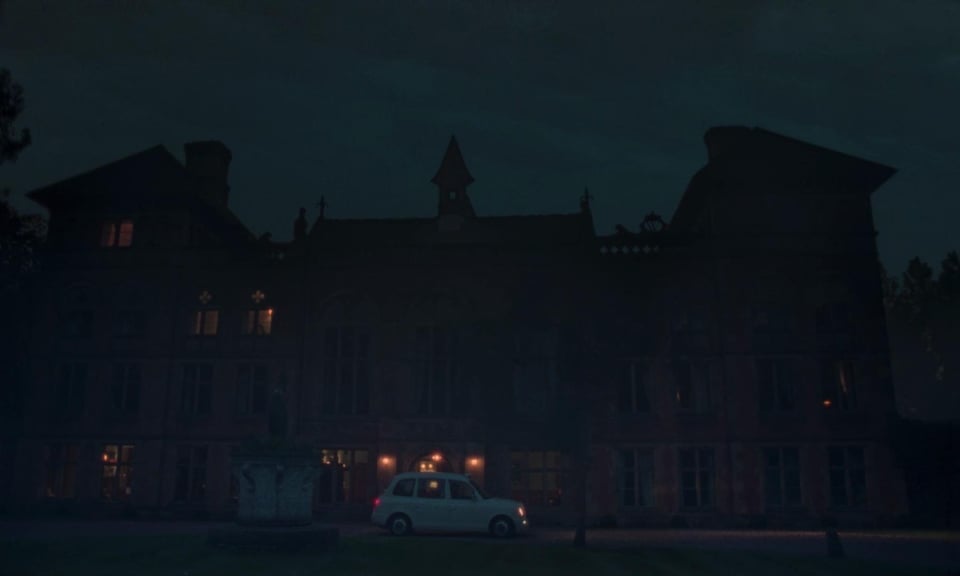 A small car idles outside of a huge old house on a misty night.