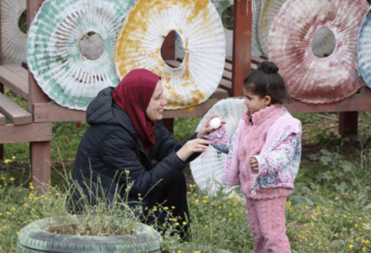 An adult with a hijab bends down in a field to show an egg to a little girl dressed in pink